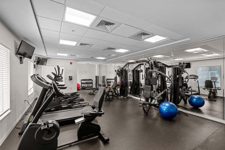 Bright fitness room with ellipticals, stationary bikes, weight equipment, blue exercise balls, and TV monitors