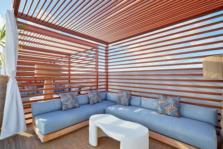 Shaded lounge area with wooden slat walls, blue sectional sofa, geometric pillows, and white curtains