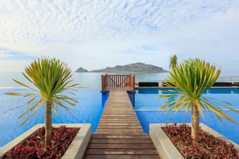 Wooden boardwalk with palm planters leading to infinity pool overlooking calm blue sea and rocky island landscape
