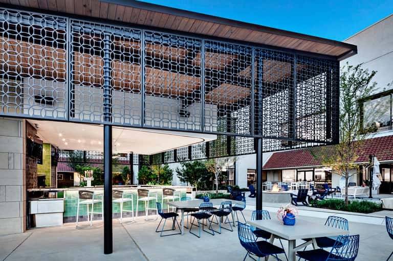 Modern outdoor courtyard with geometric patterned screens, dining tables with blue chairs, and kitchen pavilion