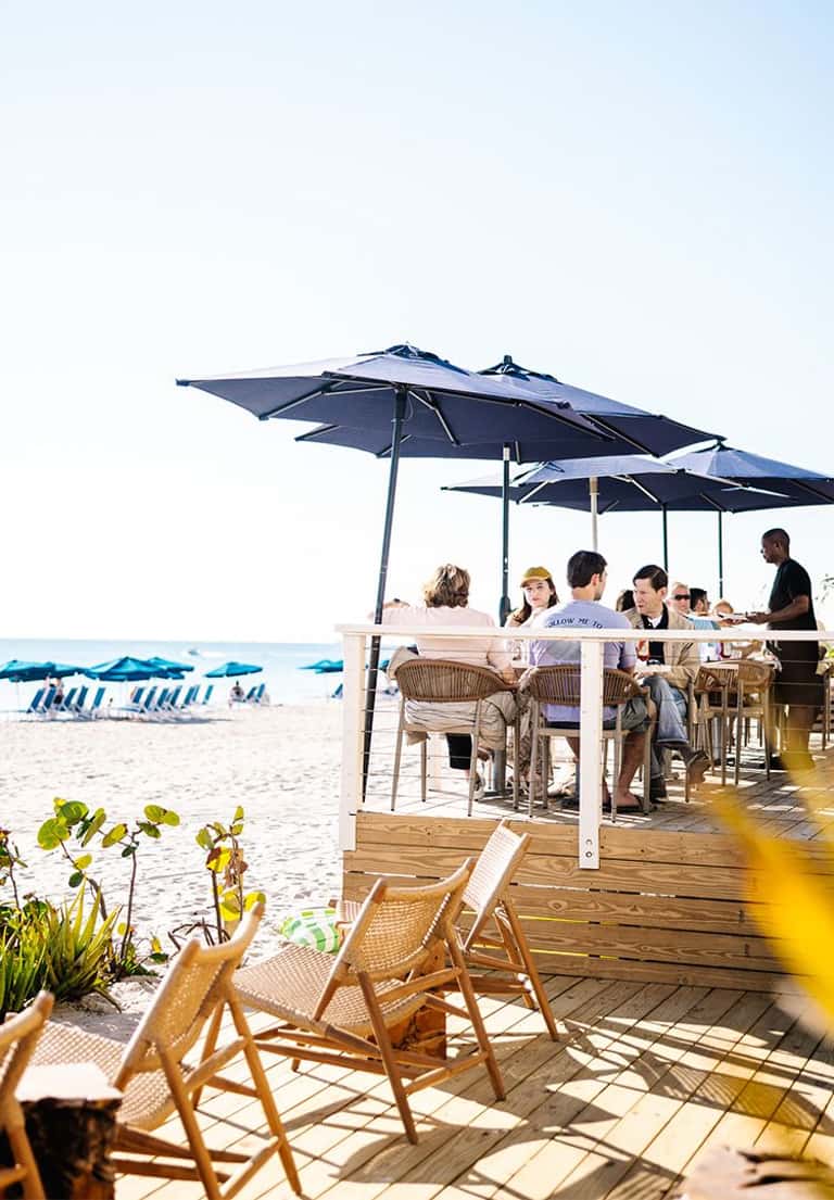 Beachfront dining area with blue umbrellas and wooden deck overlooking sandy beach and ocean