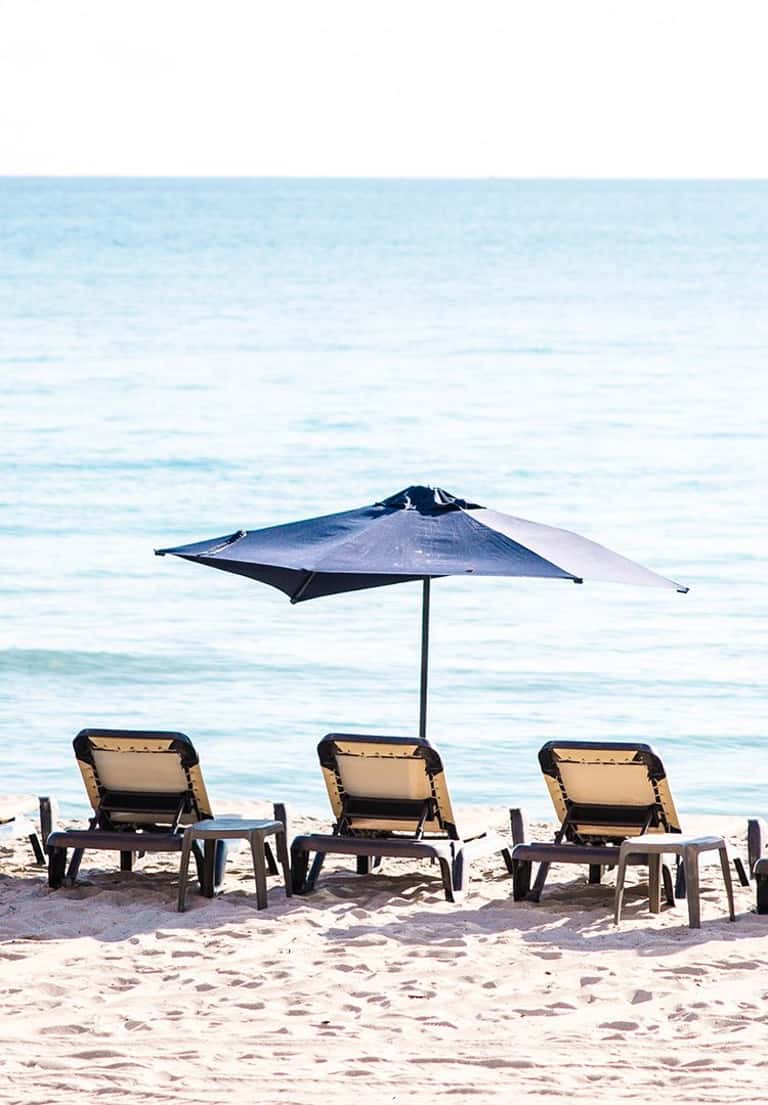 Three lounge chairs with single blue umbrella on sandy beach facing calm turquoise water