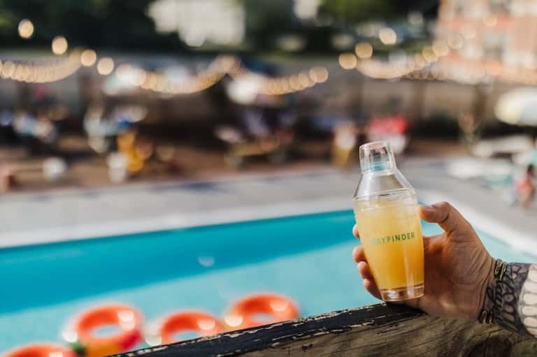Hand holding a yellow Dayfinder cocktail by the pool with inflatable floats in background