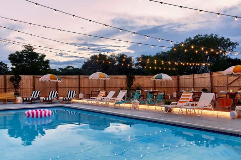 Resort pool at dusk with string lights, umbrellas, lounge chairs, and wooden fence backdrop