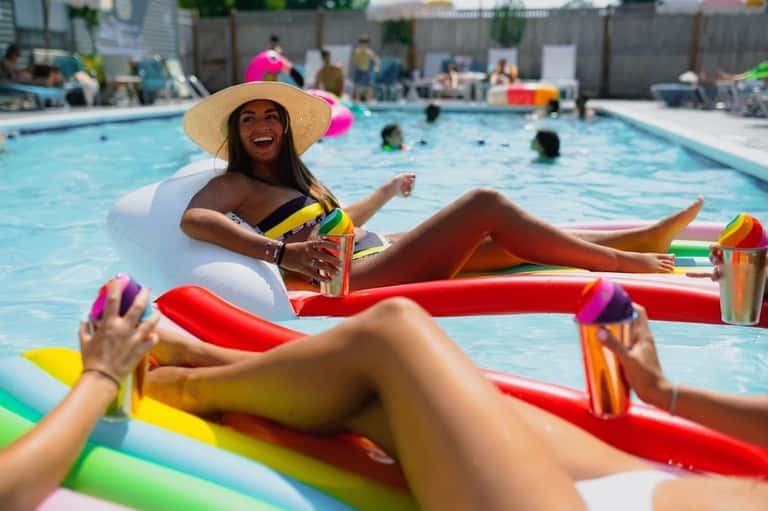 Woman relaxing on red pool float wearing sun hat, smiling with colorful drinks and other floats