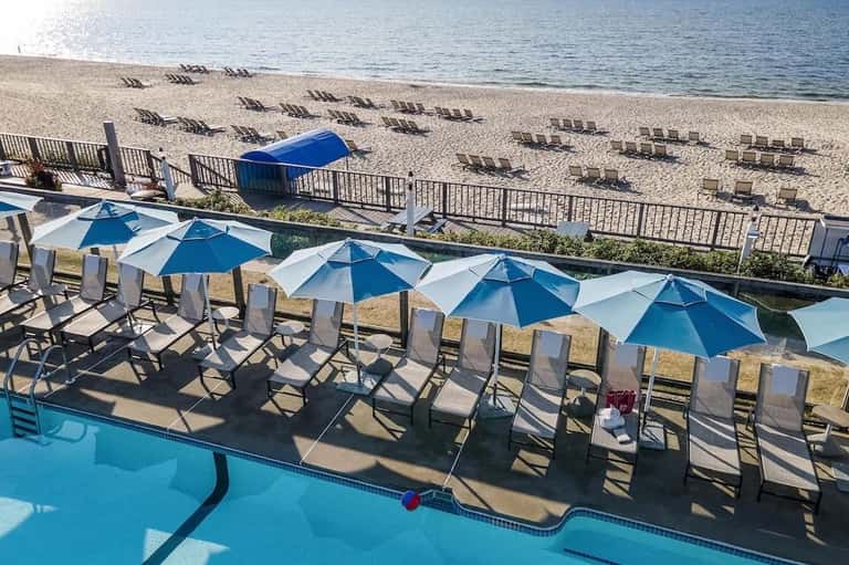 Beachfront pool area with blue umbrellas, lounge chairs, and sandy beach with sunbathers
