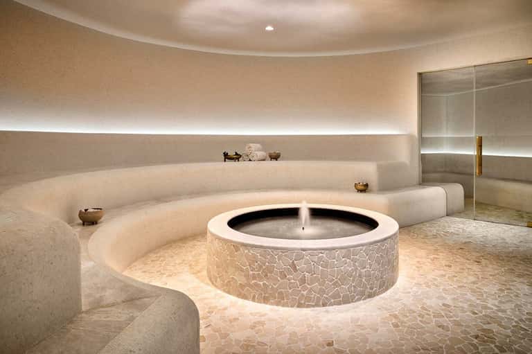 Luxurious circular spa with central fountain, curved seating, warm lighting, and glass shower enclosure