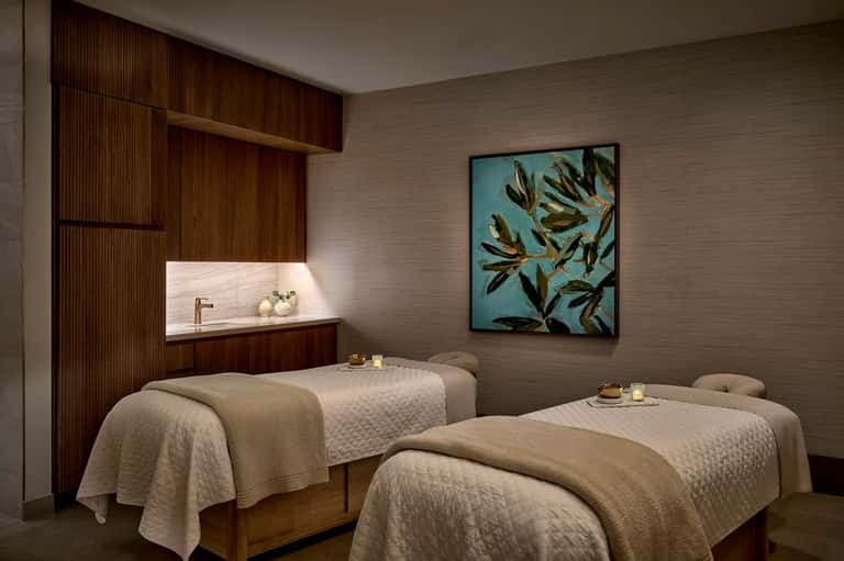Spa treatment room with two massage beds, wooden accents, artwork, and ambient lighting
