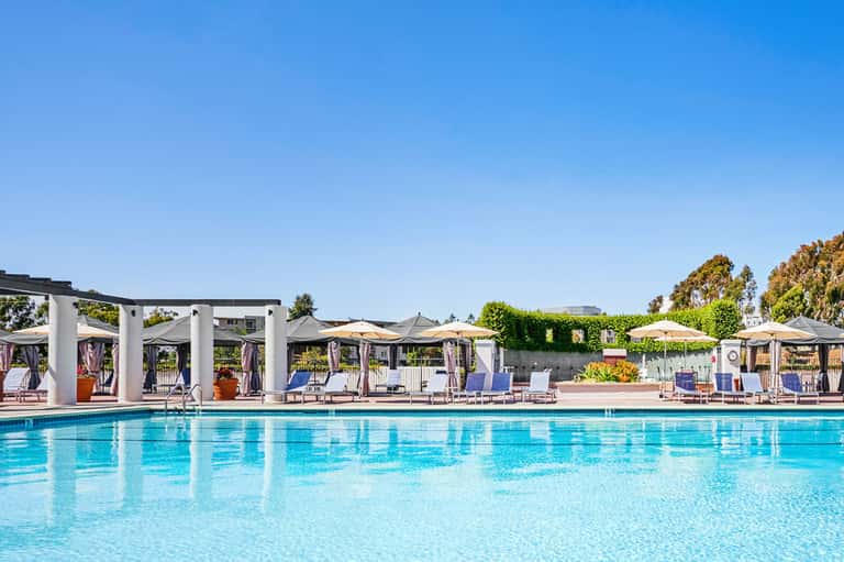 Resort outdoor pool with loungers, umbrellas, and white pergolas under clear blue sky