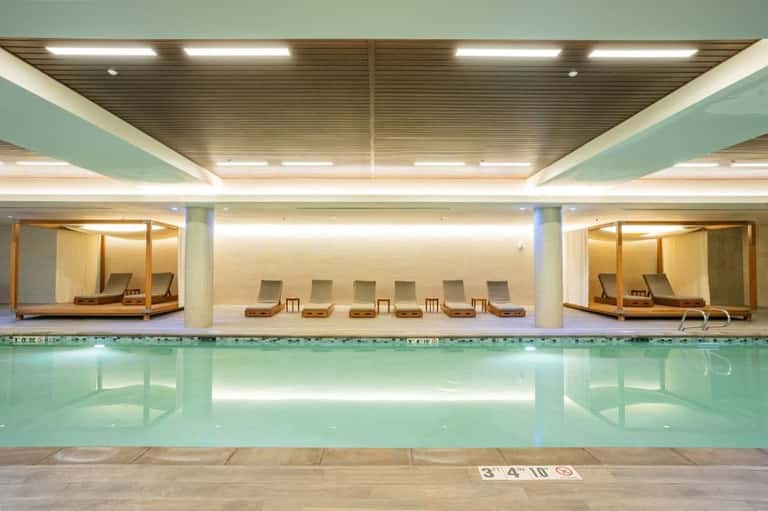 Indoor lap pool with turquoise water, lounge chairs, and relaxation alcoves with wooden canopies