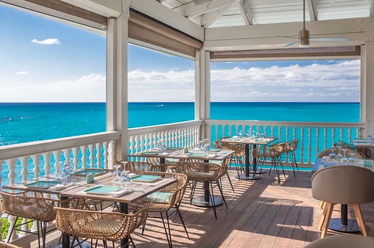Elegant beachfront dining terrace with turquoise ocean views, wicker chairs, and white railings