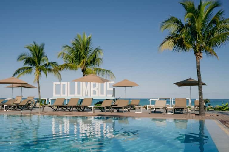 Resort pool area with Columbus signage, palm trees, lounge chairs, and umbrellas overlooking the sea