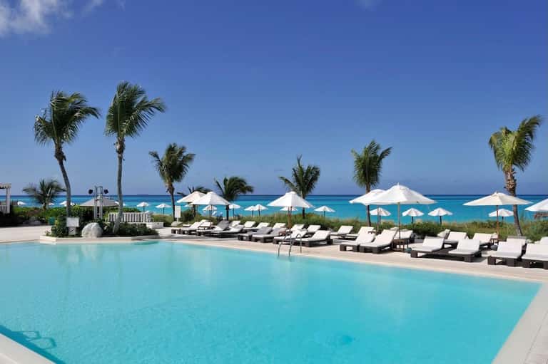Luxurious beachfront pool with white umbrellas, lounge chairs, palm trees, and turquoise ocean horizon