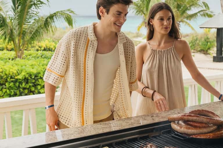 Couple grilling sausages on a beachfront deck with ocean and palm trees in background