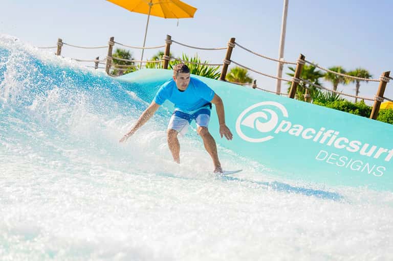 Surfer riding artificial wave on shallow water ride with palm trees and yellow umbrella