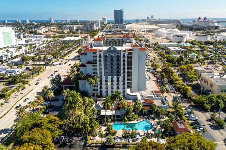 Aerial view of Spanish-style resort with pool, palm trees, and waterfront in background