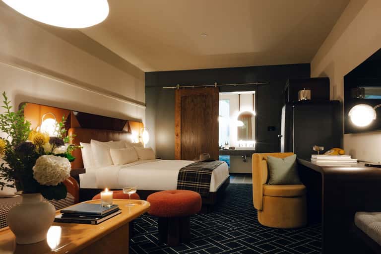 Contemporary hotel bedroom with dark accent wall, white bedding, wooden barn door, amber accent chair, and warm ambient lighting