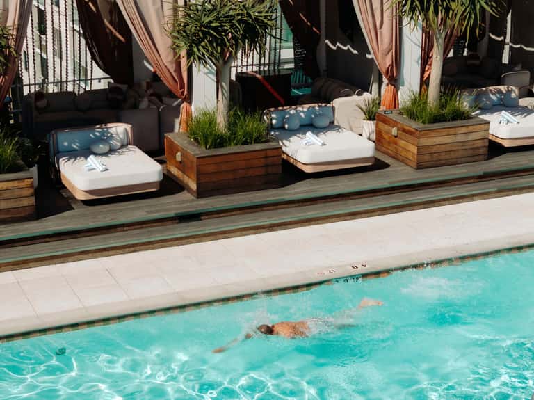 Guest swimming laps in turquoise pool with wooden cabanas, white cushions, and flowing curtains poolside