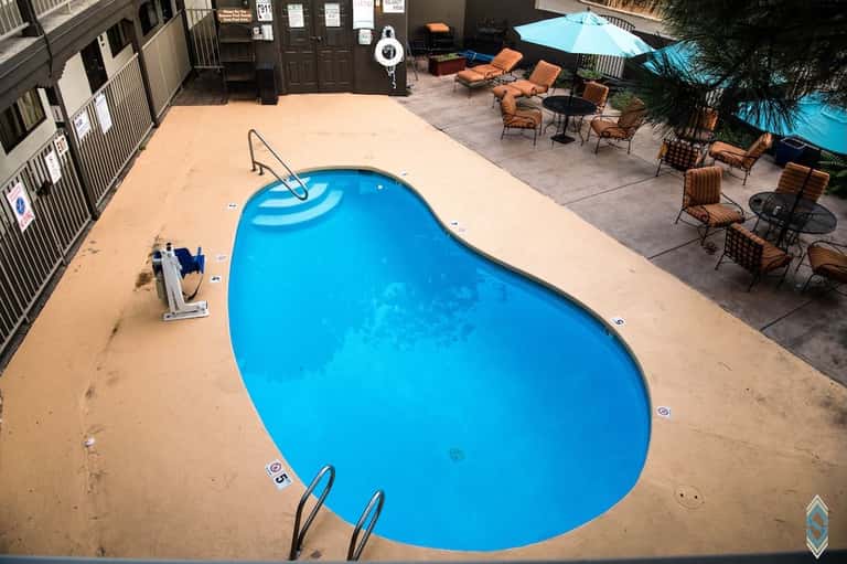 Bright blue kidney-shaped swimming pool surrounded by tan deck with lounge chairs and umbrellas under clear sky