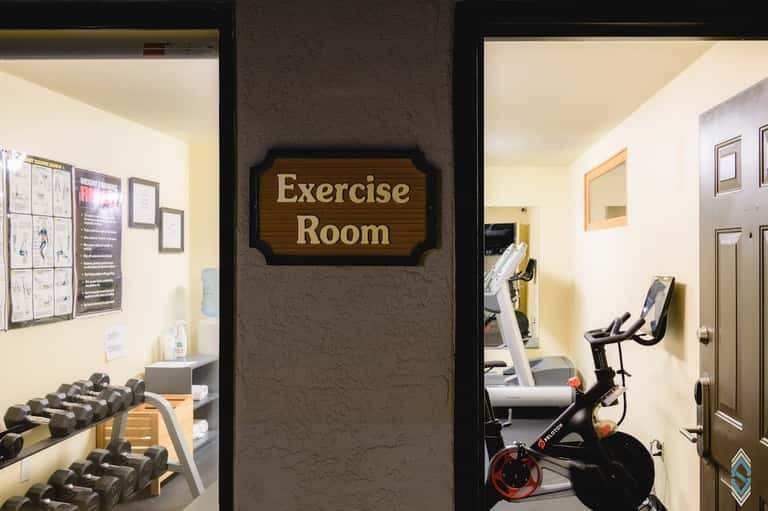Exercise room with dumbbells on rack, stationary bike, and workout equipment visible through glass doors