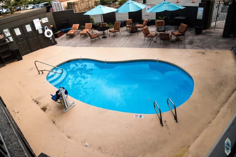 Overhead view of large blue pool with tan deck, lounge chairs, blue umbrellas, and patio area with palm trees