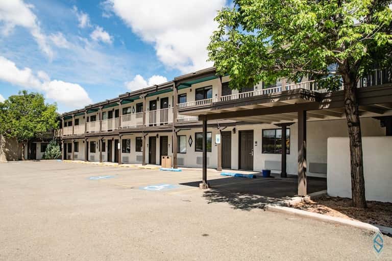 Two-story southwestern-style building with covered porch, wooden railings, and manicured courtyard with shade trees