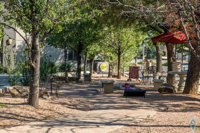Shaded outdoor recreational area with tall mature trees, cornhole game, red umbrella, and seating under tree canopy