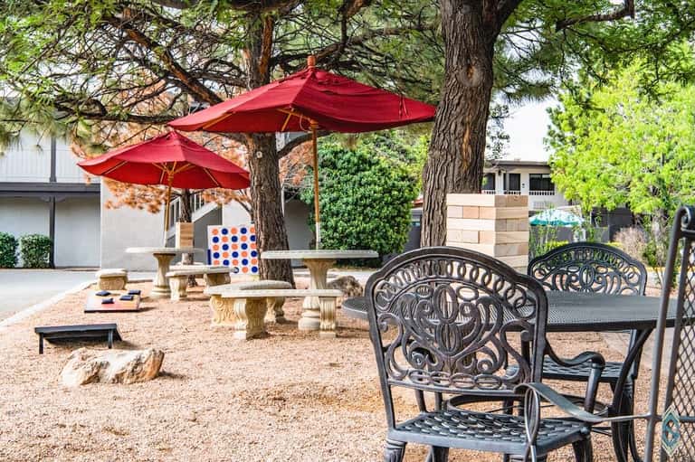 Outdoor recreation area with red umbrellas, metal chairs, stone benches, and cornhole game under trees