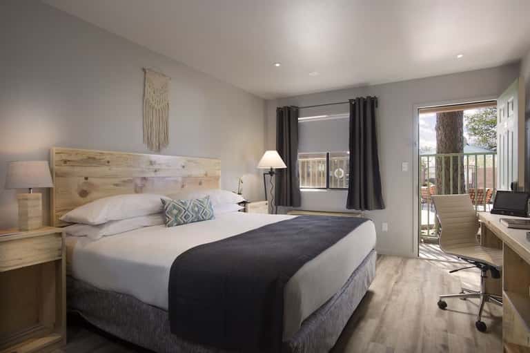 Modern hotel bedroom with queen bed, wooden headboard, desk, and glass door opening to balcony