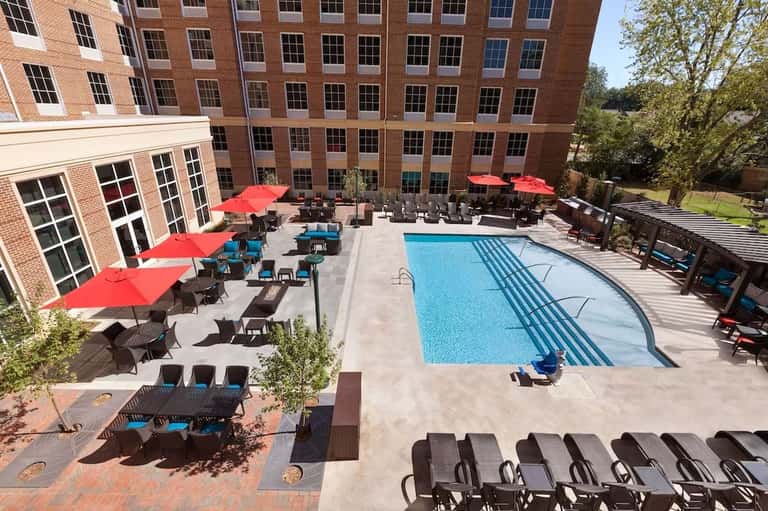 Outdoor pool area with red umbrellas, lounge chairs, and multi-story brick hotel building