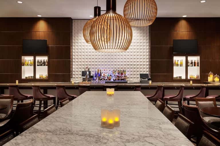 Modern hotel bar with marble countertop, wooden pendant lights, and illuminated bottle display