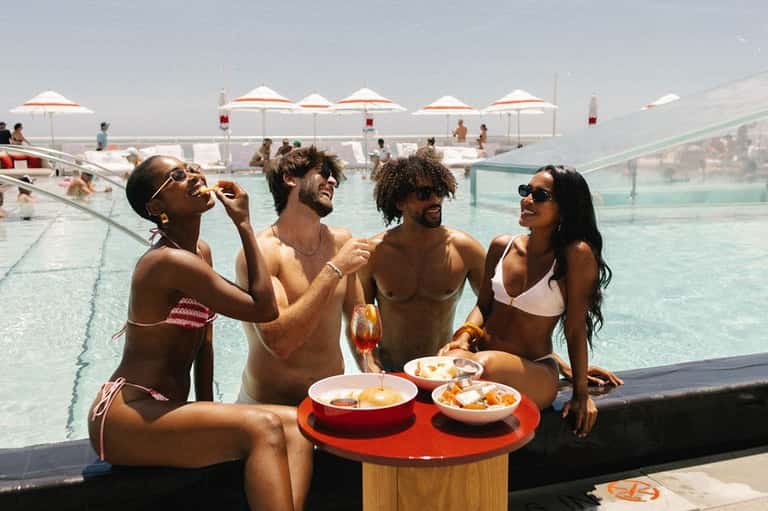 Guests enjoying pool area with white umbrellas, food service, and infinity pool views