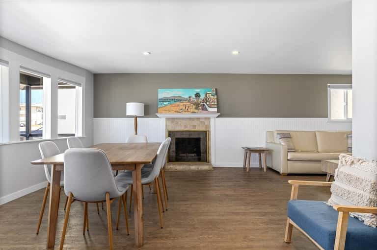 Spacious open-concept living area with dining table, fireplace, and coastal artwork, featuring white wainscoting and neutral tones