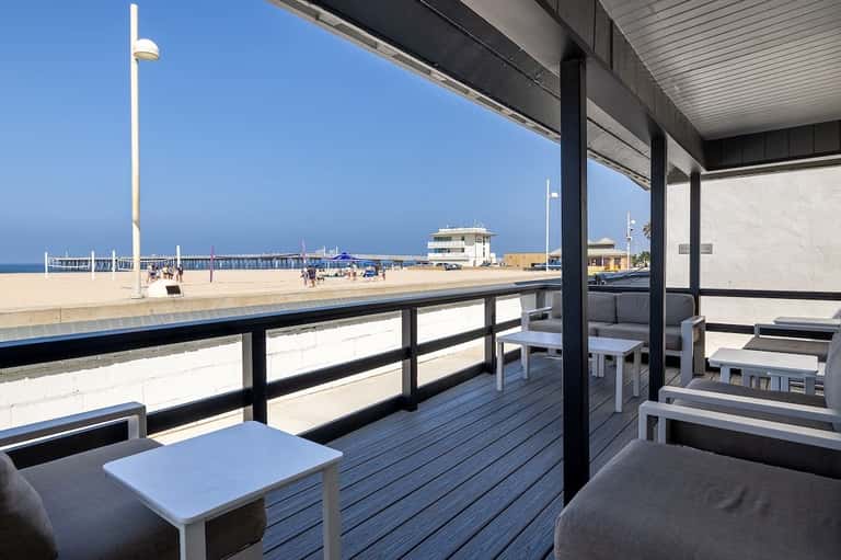 Beachfront deck with lounge seating overlooking sandy beach, pier, and ocean under clear blue sky