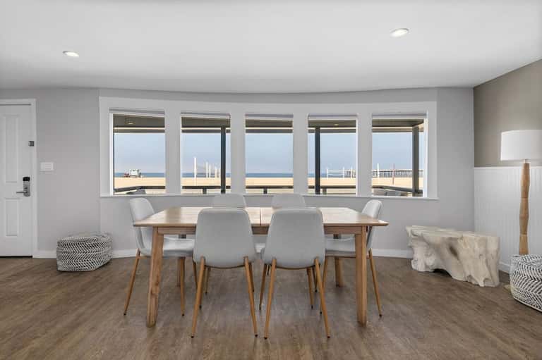 Bright dining area with wooden table, gray chairs, and expansive windows framing beach and lifeguard tower views