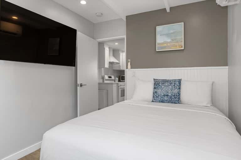 Compact bedroom with white bedding, beige and white wainscoting, and adjacent kitchenette