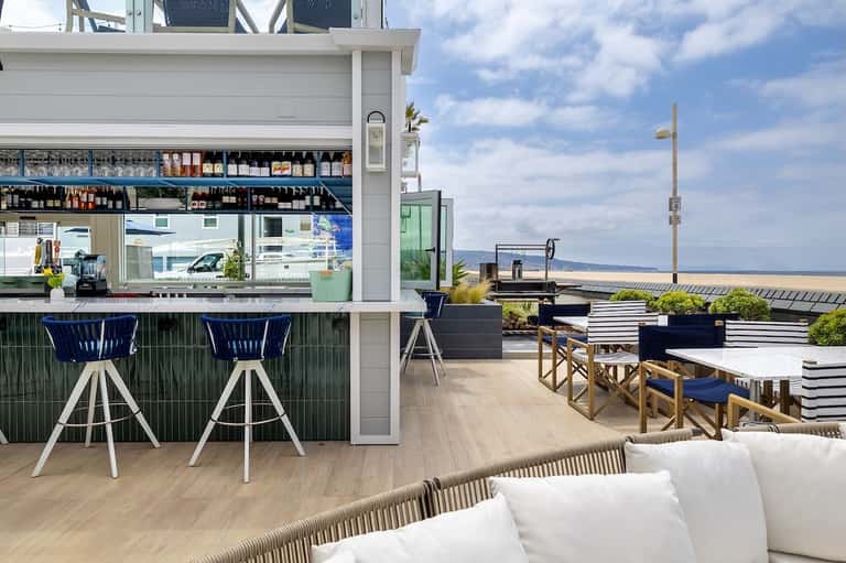 Waterfront bar and dining area with blue stools, white railings, patterned deck, and ocean backdrop