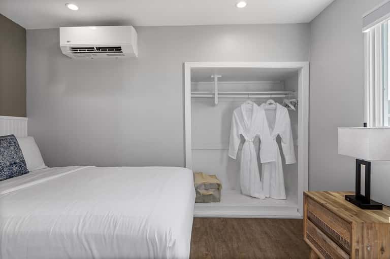 Hotel bedroom with queen bed, open walk-in closet with white robes, and air conditioning unit