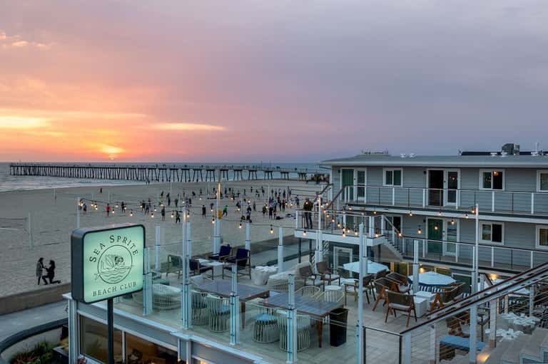 Sunset beach club scene with multi-level deck, dining areas, sunset pier view, and beachgoers below