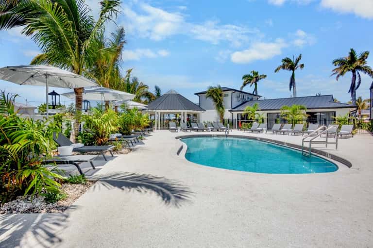 Resort pool with lounge chairs, white umbrellas, gazebo, and palm trees under blue sky