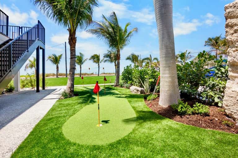 Putting green with red flag surrounded by palm trees and green grass near waterfront building