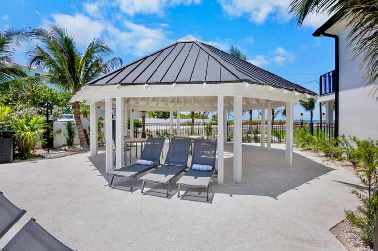 White pergola gazebo with black roof covering lounge chairs overlooking ocean waterfront