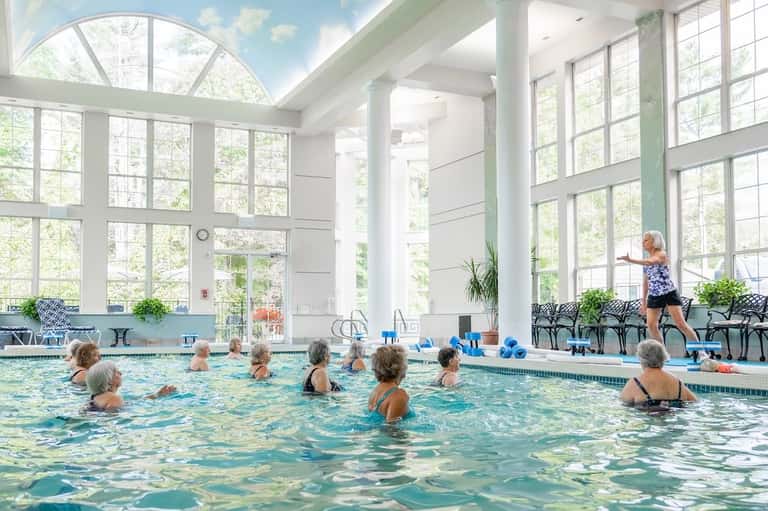 Indoor swimming pool with seniors exercising, large windows overlooking trees, instructor on deck