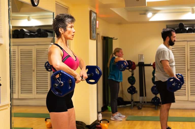 Group fitness class with people holding dumbbells in a yellow-walled studio with mirrors