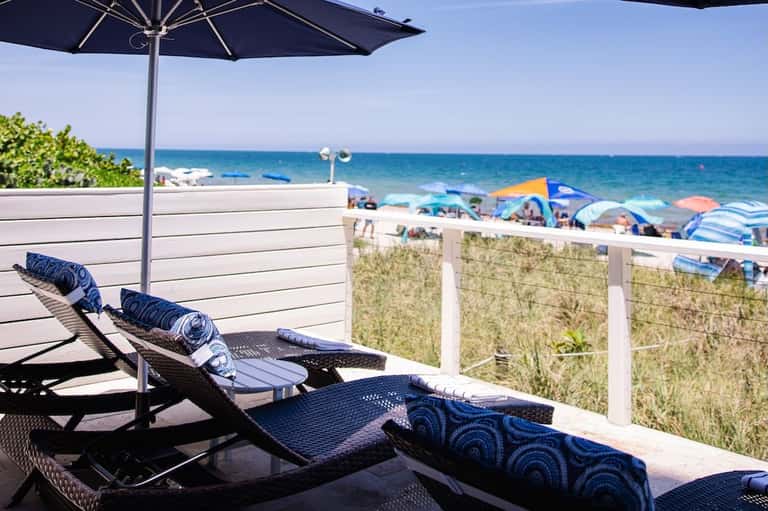 Oceanfront lounge chair with blue umbrella overlooking beach, sand dunes, and water