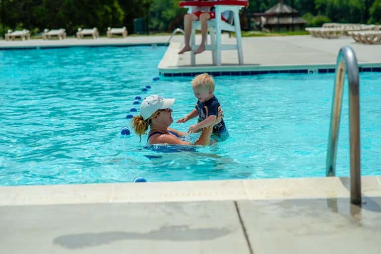 Swim instructor teaching young child in resort pool with diving board and slide visible