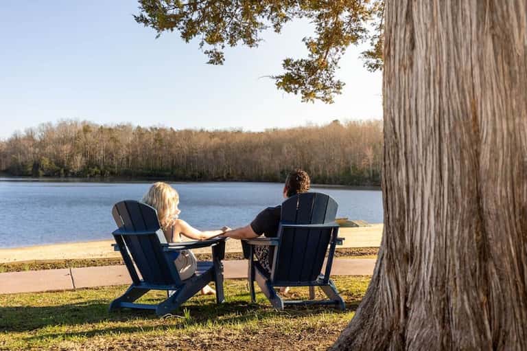 Couple in Adirondack chairs overlooking calm river with bare trees reflected in water