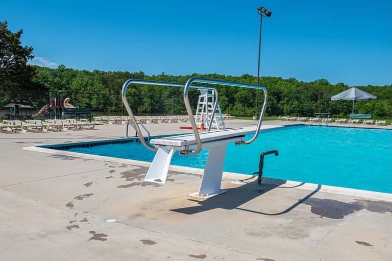 Resort swimming pool with diving board, lounge chairs, umbrellas, and wooded backdrop under clear blue sky