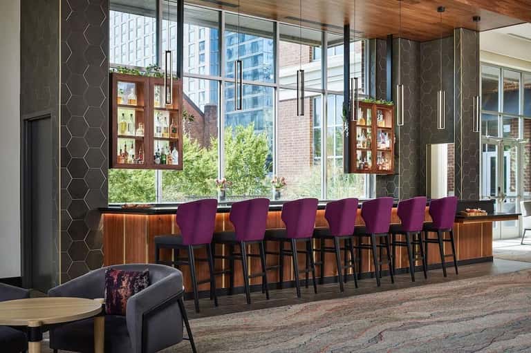 Modern hotel bar with purple bar stools, wooden cabinetry, large windows overlooking city buildings