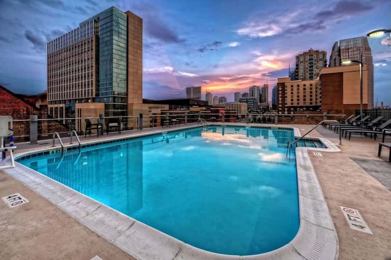 Rooftop swimming pool at dusk with city skyline, lounge chairs, and purple-pink sky